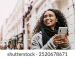 Woman holding mobile phone, walking in the city. Happy black woman holding phone while walking in the city. African-American woman walking in the city with phone. Happy black woman with phone