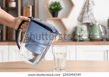 Woman holding modern filter jug near glass of water on table in kitchen