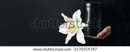 Woman holding mortuary urn and lily flower on black background with space for text, closeup
