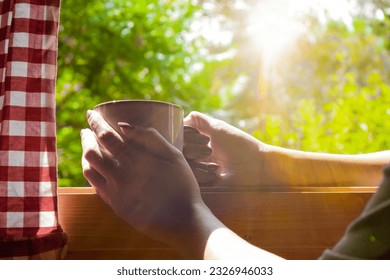 A woman holding a mug of coffee welcomes the morning sun at a window overlooking the garden. - Shutterstock ID 2326946033