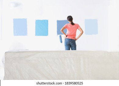 Woman holding a paint roller is looking at samples of blue paint on the living room wall to decide the right shade.