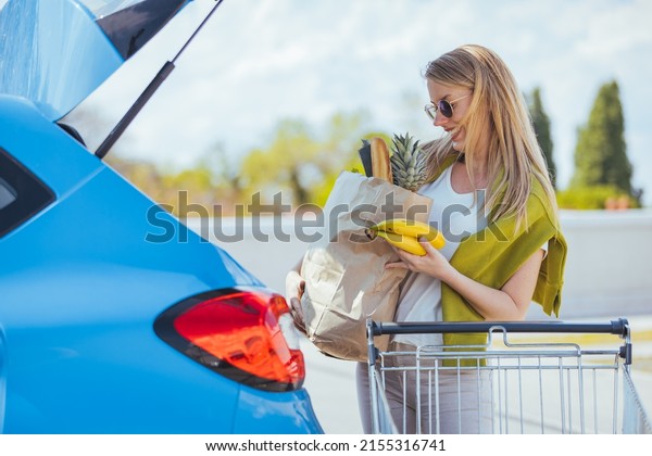Woman holding paper bags with food products.\
Young woman putting package with groceries and vegetables into car\
trunk. Attractive caucasian female shopping in mall or grocery\
store. Zero waste concept