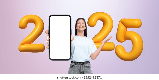 A woman is holding a phone mockup with blank screen, smiling, with large golden 2025 balloons on a gradient background, concept of New Year celebration
