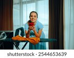 
Woman Holding Phone While Doing her Daily Chores at Home. Happy housewife spending time online instead of ironing clothing
