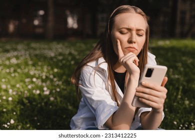 Woman holding a phone while holding her hand on chin. Expressive student thinking outside in campus - Woman in doubt with smartphone in her palm, think about reading information, search solution. - Shutterstock ID 2313412225
