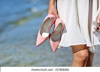 Woman Holding Pink Stiletto Heels On The Beach Walking In White Dress