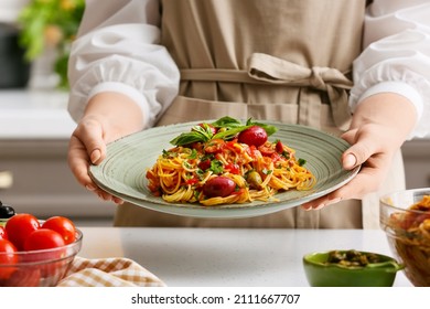 Woman holding plate with tasty Pasta Puttanesca at table in kitchen, closeup