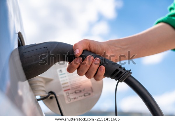 Woman holding plug of electric car\
charging station on a background of blue sky. Close\
up