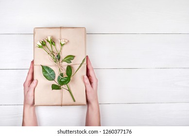 Woman Holding Present Box With Rose Flowers On Wooden Table, Copy Space. Gift In Girl's Hands, Free Space. Holiday Background For Womens Day, Mothers Day. Flat Lay, Top View. Gift Wrapping Background