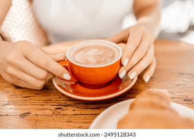 A woman is holding a red coffee cup with a white saucer. The woman is sitting at a table with a plate of croissants in front of her. - Powered by Shutterstock