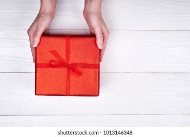 Woman Holding Red Present Box On White Wooden Table Background, Free Space. Close Up Of Gift Box In Girl's Hands, Copy Space. Holiday Background. Flat Lay, Top View. Gift Wrapping Background