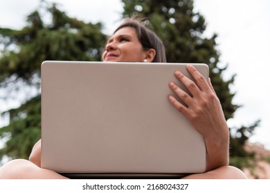 Woman holding the screen of her laptop with one hand while looking at infinity. Image from below of a smiling young girl who is using her computer outdoors on a cloudy day. Technology life concept.