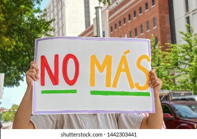 Woman Holding Up Sign That Says No Mas - Spanish For No More - In Front Of Her Face With Tall Buildings And Cars In The Background And Another Protest Sign Seen To The Side