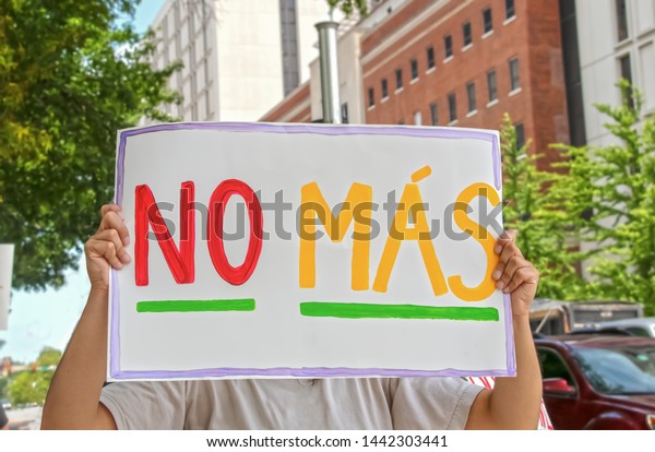 Woman holding up\
sign that says No Mas - Spanish for No More - in front of her face\
with tall buildings and cars in the background and another protest\
sign seen to the side