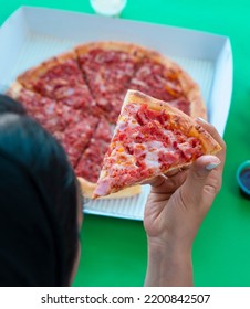 A Woman Is Holding A Slice Of Pizza With Her Hand In Front Of Her Mouth, A Pizza Made Of Salami, Beacon And Cheese In A Box Is In Front Of Her On A Green Background.