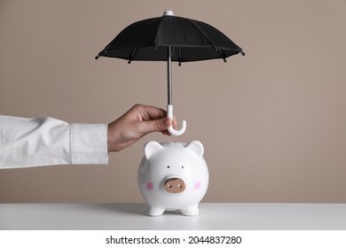 Woman holding small umbrella over piggy bank against beige background, closeup