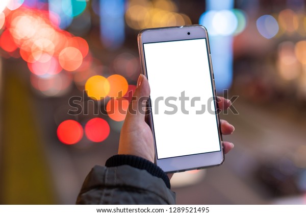 Woman holding a\
smartphone with a blank white screen at night with lights from car\
traffic in the background