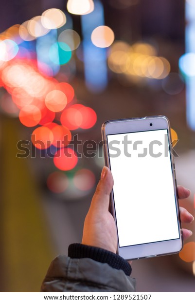 Woman holding a\
smartphone with a blank white screen at night with lights from car\
traffic in the background