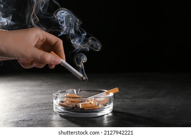 Woman holding smoldering cigarette over glass ashtray at grey table against black background, closeup