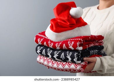 Woman holding stack of different Christmas sweaters and Santa hat on grey background, closeup. Space for text - Powered by Shutterstock