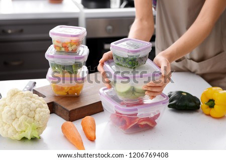Woman holding stack of plastic containers with fresh vegetables for freezing at table in kitchen