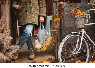 Woman Holding String Bag With Autumn Seasonal Vegetables While Standing At Door Of Old Wooden House