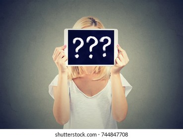 Woman holding tablet pc with question marks hiding her face isolated on gray background.