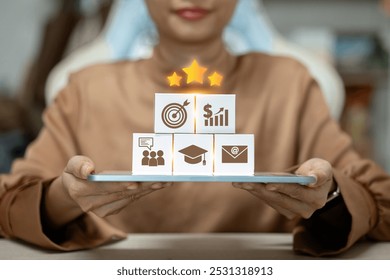 A woman is holding a tablet with a stack of blocks on it. The blocks have different symbols on them, including a star, a graduation cap, and a dollar sign - Powered by Shutterstock