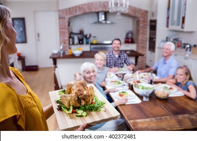 Woman Holding A Tray Of Roasted Turkey And Family Dining In Background