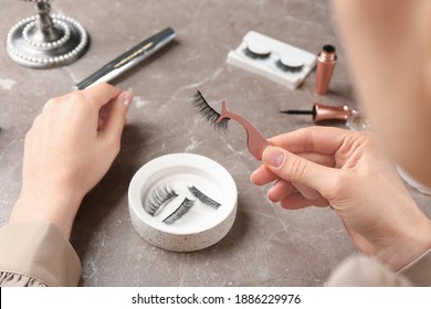 Woman holding tweezers with magnetic eyelashes at grey table, closeup