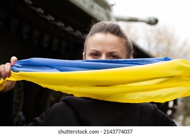 Woman Holding Ukrainian Flag, With Battle Tank In Background. Russia Invasion, War In Ukraine, Crisis Concept. Girl Standing Next To Military Armored Fighting Vehicle.