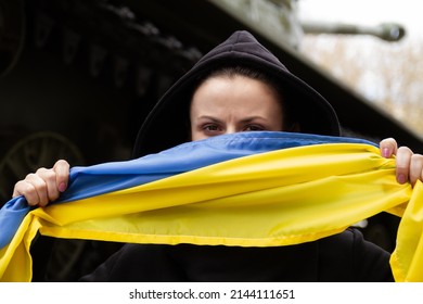 Woman Holding Ukrainian Flag, With Battle Tank In Background. Russia Invasion, War In Ukraine, Crisis Concept. Girl Standing Next To Military Armored Fighting Vehicle.