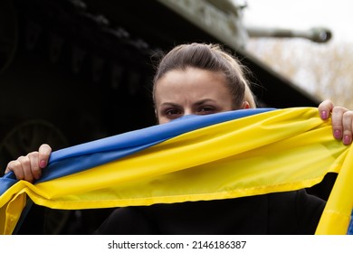 Woman Holding Ukrainian Flag, With Battle Tank In Background. Russia Invasion, War In Ukraine, Crisis Concept. Girl Standing Next To Military Armored Fighting Vehicle.