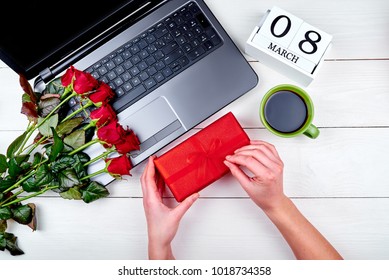 Woman Holding And Unwrapping Gift Box. Womens Day Background With Bouquet Of Red Roses, Gift Box, Calendar With Date March 8, Cup Of Coffe And Open Laptop Computer, Copy Space. Top View, Flat Lay