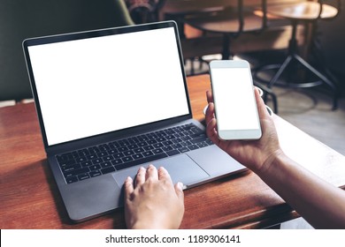Eine Frau, die ein Mobiltelefon mit weißem Bildschirm und einem Laptop auf Holztisch im Cafeteria hält und benutzt, mit einem Mock-up-Handy