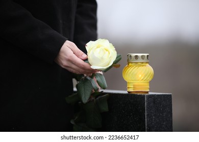Woman holding white rose near black granite tombstone with candle outdoors, closeup. Funeral ceremony