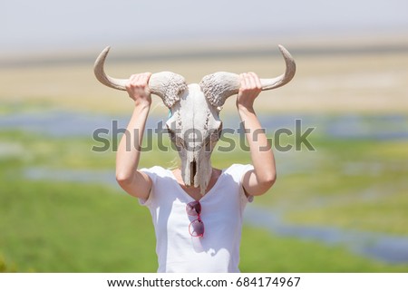 Woman holding a white wildebeest skull wearing it like a mask in nature on african wildlife safari, Amboseli national park, Kenya.