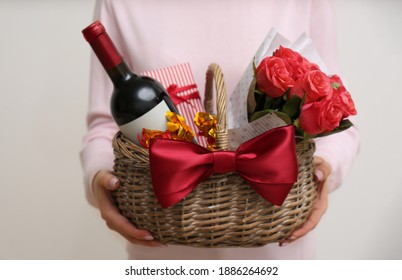 Woman holding wicker basket with gift, bouquet and wine on light grey background, closeup - Shutterstock ID 1886264692