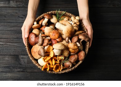 Woman holding wicker bowl with different wild mushrooms at black wooden table, top view