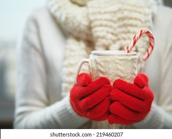 Woman holding winter cup close up on light background. Woman hands in woolen red gloves holding a cozy mug with hot cocoa, tea or coffee and a candy cane. Winter and Christmas time concept.