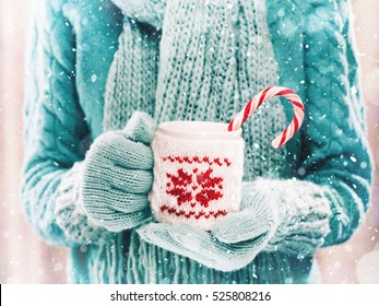 Woman holding winter cup close up on light background with snowfall. Woman hands in teal gloves holding a cozy mug with hot cocoa, tea or coffee and a candy cane. Winter and Christmas time concept.