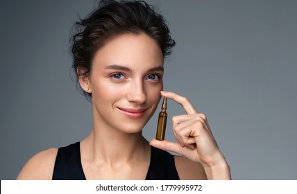 Woman Holds Ampoule With Serum For Hair Or Skin Care. Photo Of Attractive Woman With Perfect Makeup On Gray Background. Beauty Concept