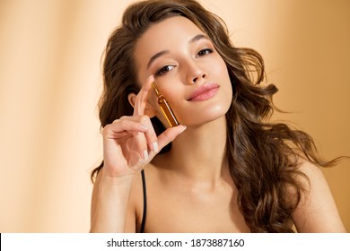 Woman Holds Ampoule With Serum For Hair Or Skin Care. Photo Of Attractive Woman With Perfect Makeup On Beige Background. Beauty Concept