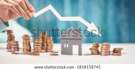 Woman holds an arrow down over model of the house and stack of coins. Concept of the crisis in the real estate market.