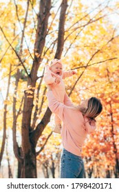 A woman holds a baby girl and lifts her up above her head. Mom and daughter have fun in nature in autumn park