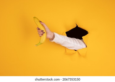 A woman holds a banana in her hand, inserted through a hole in torn yellow paper