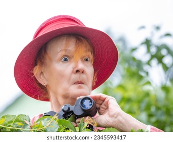 A woman holds binoculars and wears a hat. She watches secretly and intensely over a green hedge. - Powered by Shutterstock