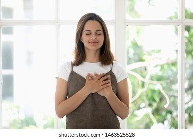 Woman holds both hands on heart symbol of gratitude closed eyes enjoy moment of love and appreciations, girl stands pose near panoramic window behind which seen green foliage as symbol of life concept - Powered by Shutterstock