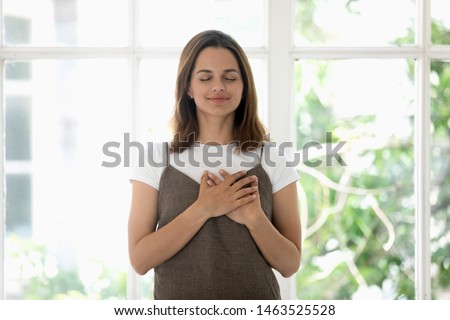 Woman holds both hands on heart symbol of gratitude closed eyes enjoy moment of love and appreciations, girl stands pose near panoramic window behind which seen green foliage as symbol of life concept
