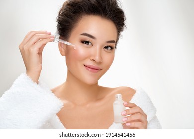 Woman holds bottle with serum for skin care. Photo of asian woman after shower on white background. Beauty and skin care concept
